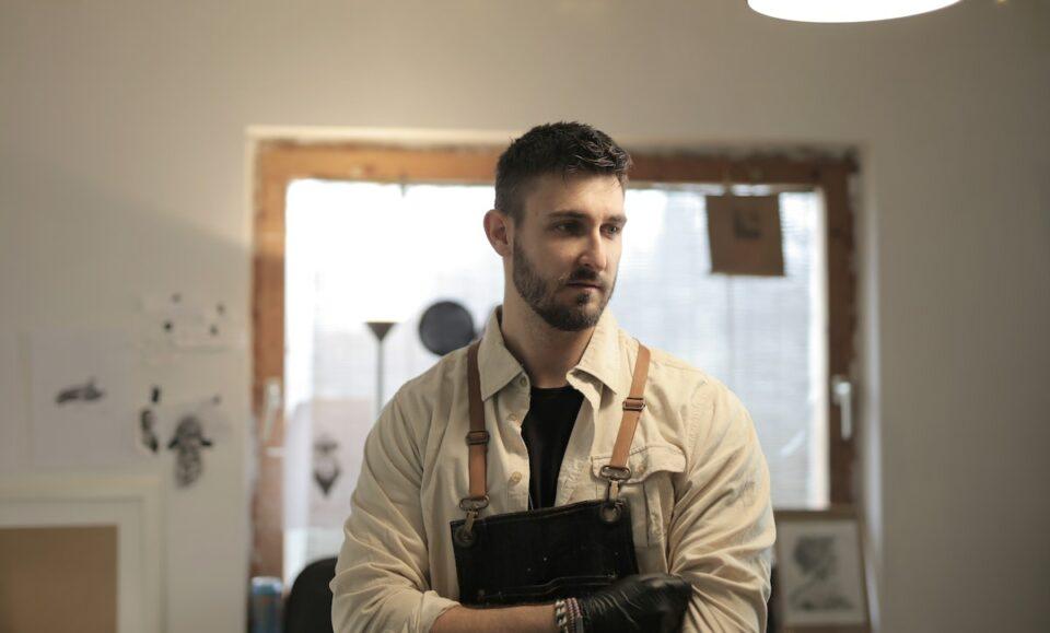 Confident young bearded craftsman in apron standing in modern workshop